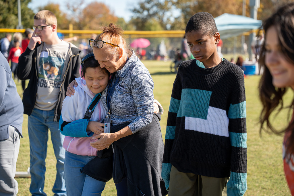 A woman in a grey sweatshirt gently hugs a girl with her arms wrapped around her. A teenage boy in a black and teal sweater stands to the right, and other people are visible in the background on the sunny field.