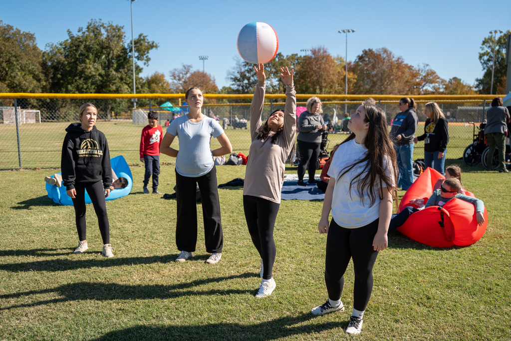 Three high school-aged volunteers or participants stand on a sunny grassy field. The person in the center is reaching up to tap a large beach ball in the air. Other people relax on inflatable chairs and watch games in the background.
