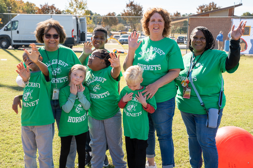 A group of six elementary school-aged children and three adults, all wearing bright green "Hillcrest Hornets" shirts, smile and wave at the camera on a sunny day.