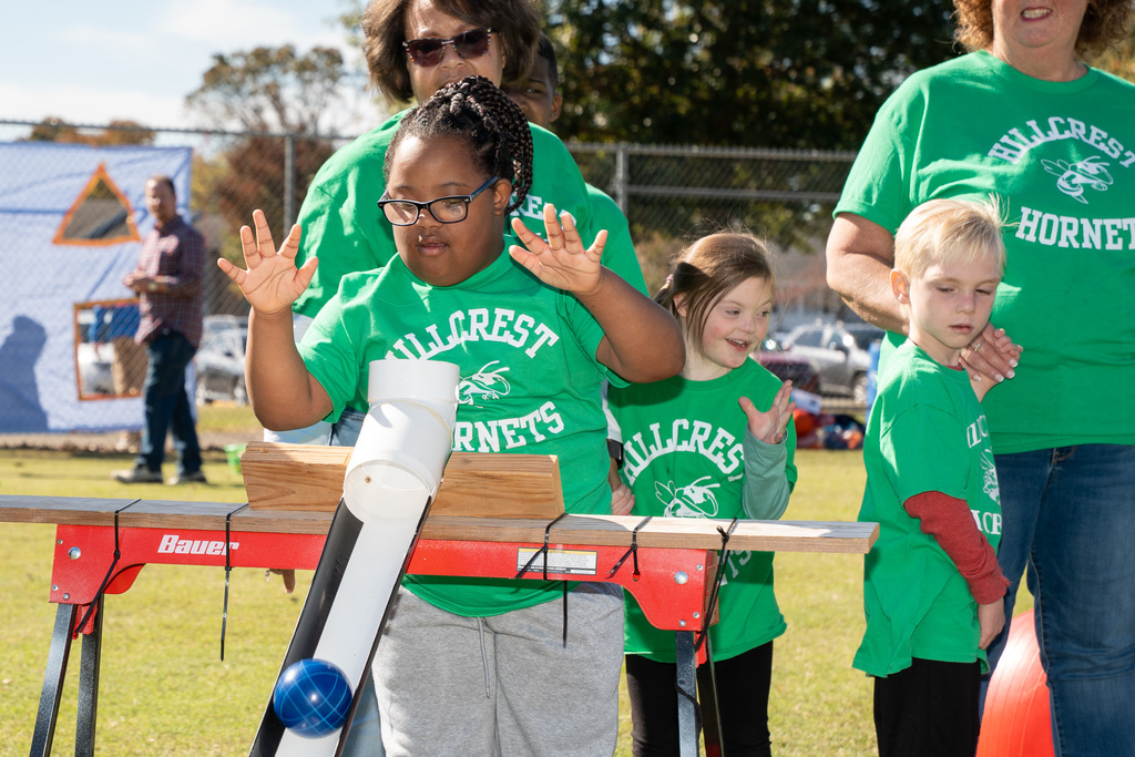 A girl in a green "Hillcrest Hornets" shirt and glasses uses a ramp device to roll a blue ball for a game, raising her hands in excitement. Another girl and several adults stand behind her, smiling.
