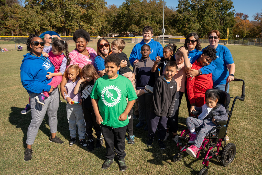 A large group of children and adult staff/volunteers pose together on a sunny grassy field, likely at a park. Several participants are wearing blue shirts. One boy in the front is wearing a green shirt, and a child sits in a pink and black wheelchair to the right.