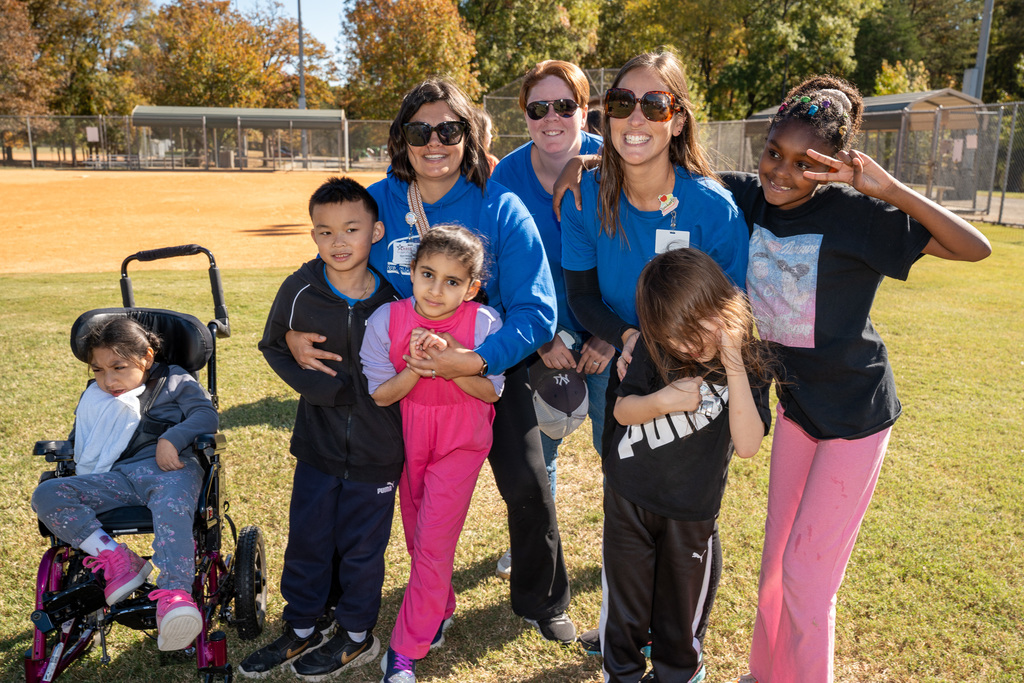 A group of five children and three adult staff/volunteers in blue shirts pose closely together, smiling on a sunny day. One girl is seated in a wheelchair on the far left.