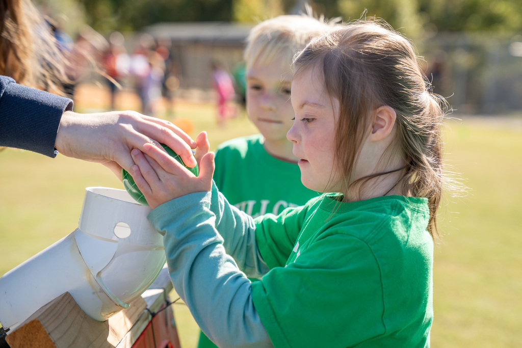 A close-up of a young girl in a green shirt carefully placing a green ball into a white plastic ramp to roll it for a game. A boy stands behind her.
