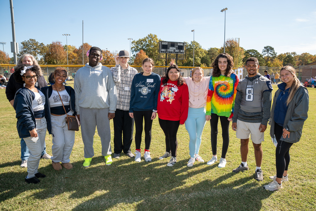 A group of about ten high school-aged students, including participants and volunteers, stand together in a line on a grassy field, posing for a group photo on a sunny day.