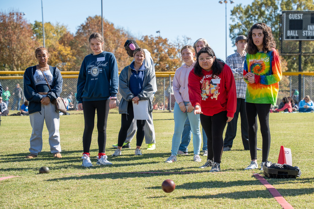A group of high school students stands on the field, focused on a game of bocce ball. One girl in a red sweatshirt is poised to roll a ball, while others watch the balls already on the ground.