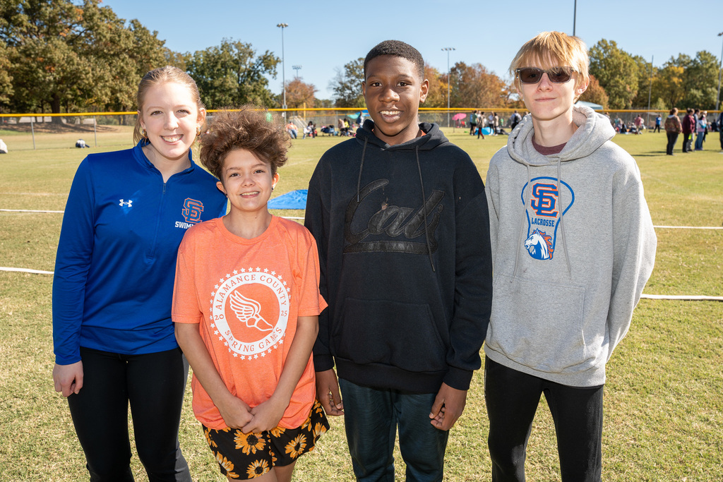 A group of four students and a volunteer stand side-by-side on a grassy field. The students are wearing a bright orange t-shirt, a black hoodie, and a grey lacrosse hoodie. The volunteer is in a blue long-sleeve athletic shirt.