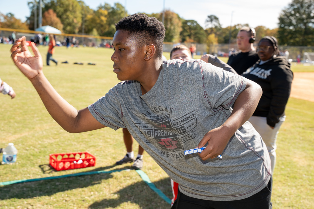 A close-up side view of a teenage boy in a grey t-shirt preparing to throw a small object with his right hand during a game on the grass. A red basket of balls is visible on the ground nearby.