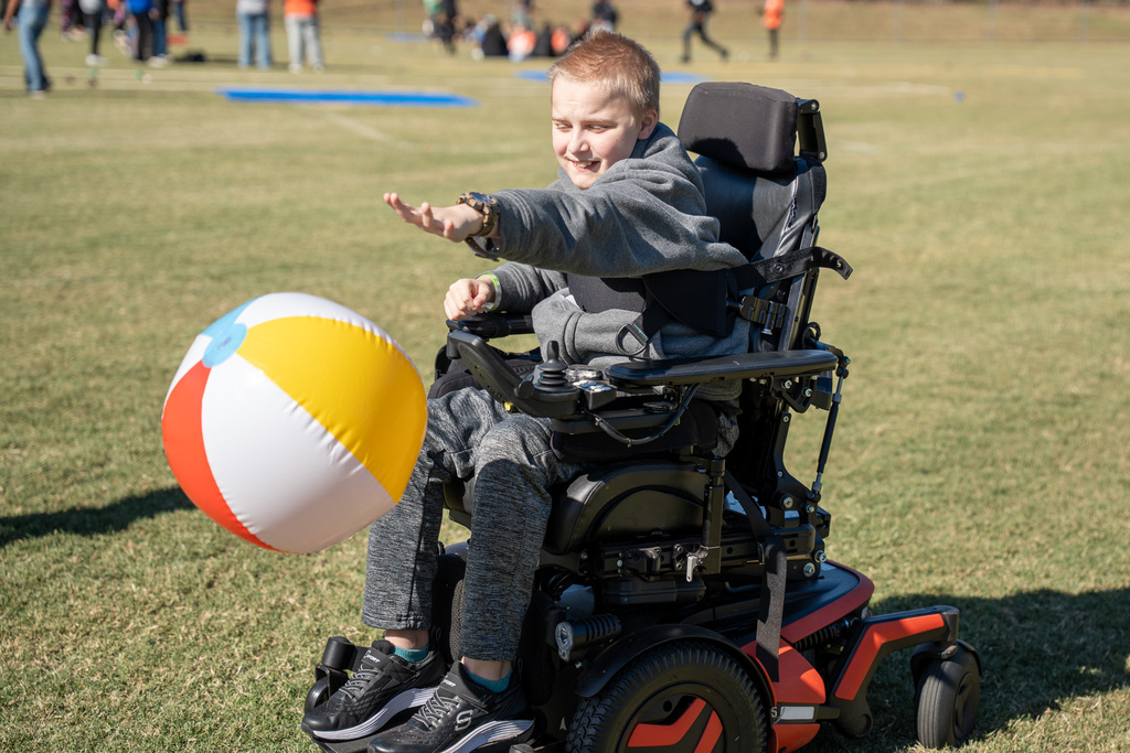 A teenage boy in a motorized wheelchair smiles as he hits a large, multi-colored beach ball forward with his right hand during an outdoor game on a grassy field.