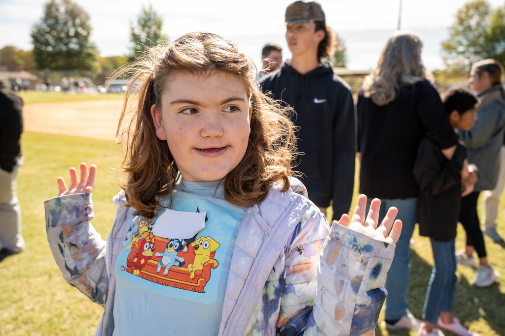 A close-up portrait of a smiling girl with long brown hair and a light blue shirt featuring a cartoon dog graphic. She holds her hands up with open palms on a sunny day.