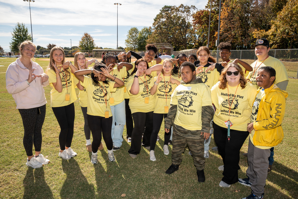 A large group of high school students, most wearing matching yellow shirts that say "United We Play, United We Win," pose for a spirited group photo on a grassy field, making silly faces and hand gestures.