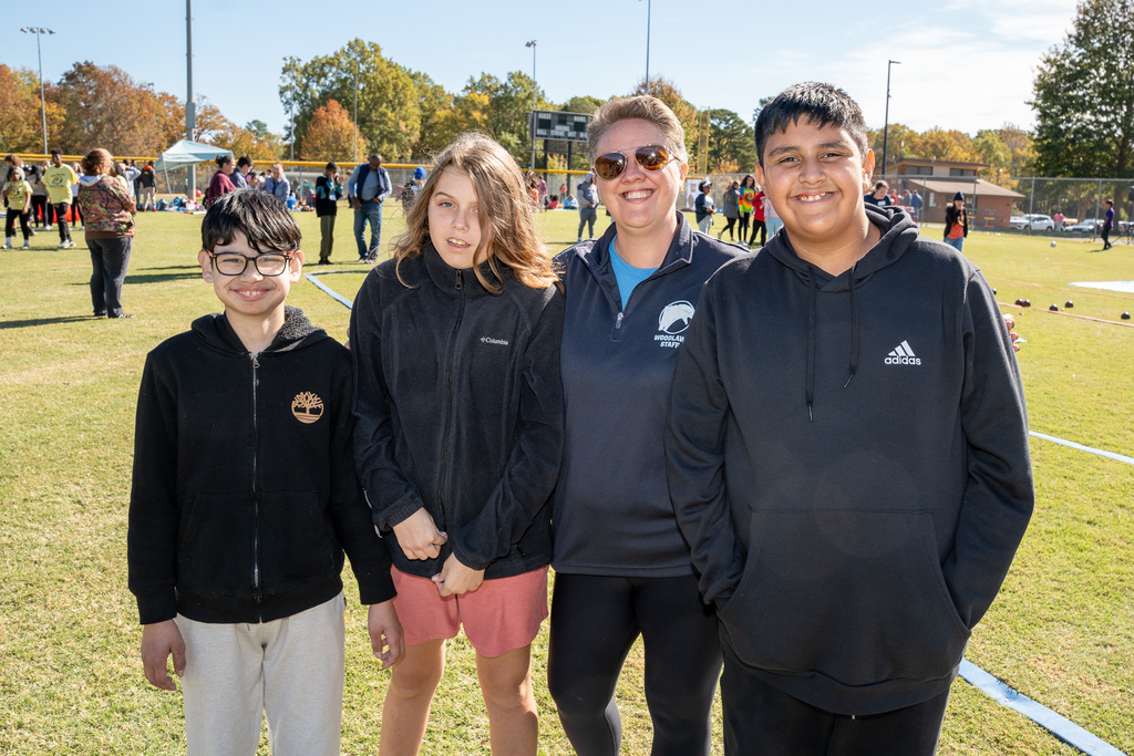 A group of four people—three students and one adult woman—stand together and smile for a close-up photo on the sunny field. The adult wears sunglasses and a blue polo shirt.