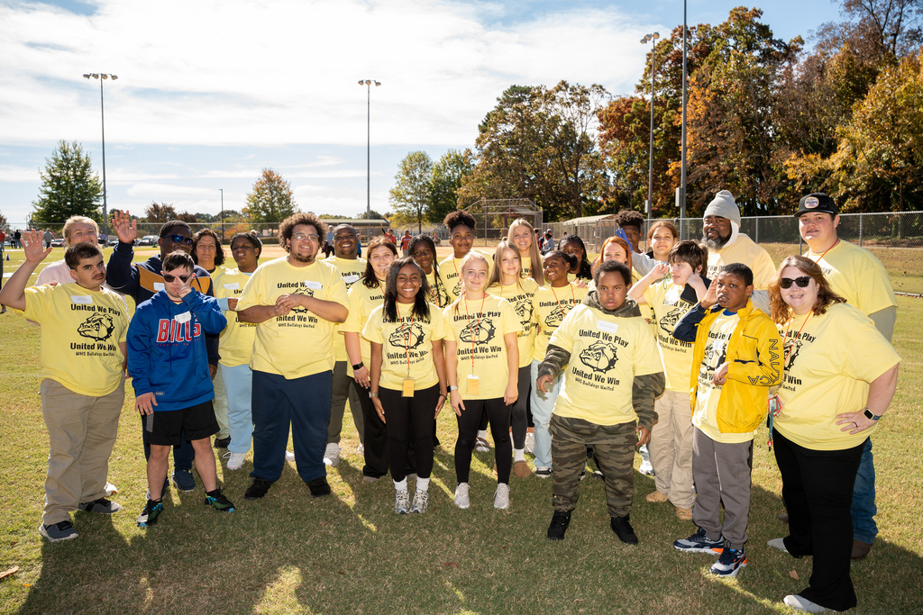 A large, diverse group of students and adult staff/volunteers, most wearing matching yellow "United We Play, United We Win" shirts, stand on a sunny grassy field and wave or smile for a group photo.
