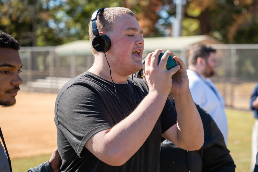 A large group of high school students and volunteers stand on a sunny grassy field surrounding a seated student in a wheelchair who is holding a beach ball. Everyone is smiling for the group photo.