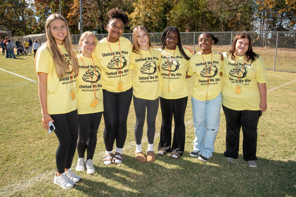 A group of seven high school girls, all wearing matching yellow "United We Play, United We Win" shirts, stand close together on a grassy field and smile for a group photo.