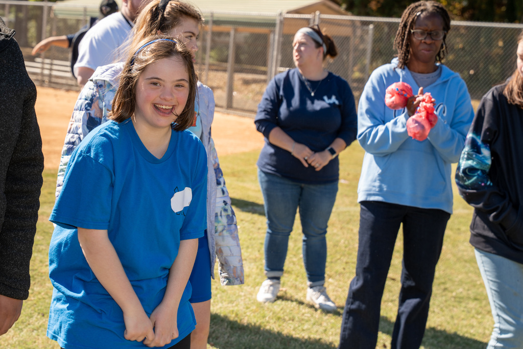 A teenage girl in a bright blue t-shirt smiles at the camera with her hands clasped in front of her. Several adults and teens stand in the background on the sunny field.