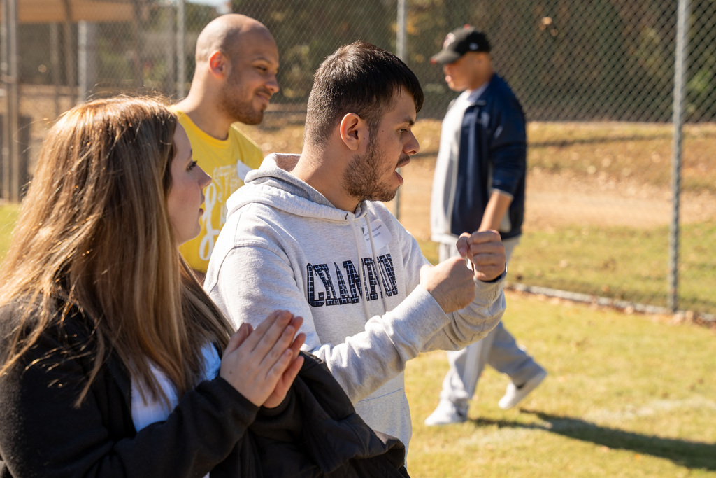 A side profile of a young man in a grey "Champion" hoodie celebrating or cheering with a clenched fist. A female volunteer in a black sweater is clapping beside him.