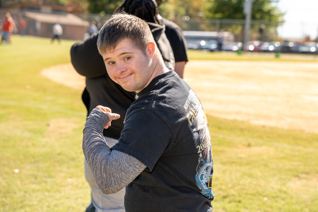 g	A young man in a black t-shirt smiles over his shoulder at the camera, pointing back at his bicep. The photo is taken on a sunny grassy field with other people and a baseball diamond in the distance.