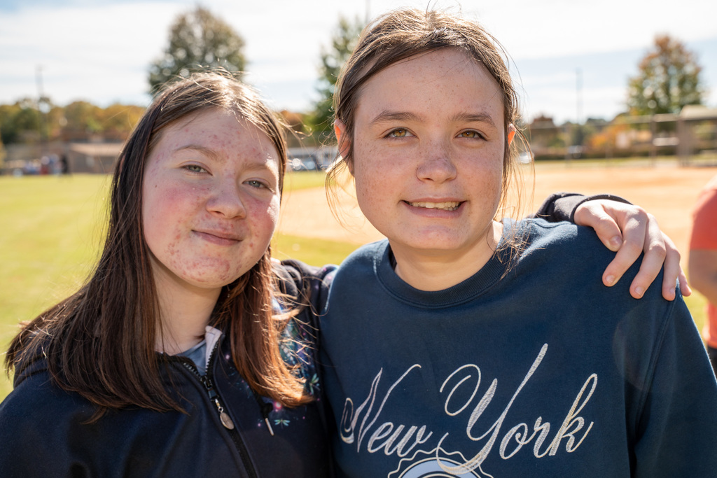 A close-up portrait of two teenage girls standing side-by-side on the field, smiling at the camera with their arms around each other. One girl wears a navy blue sweatshirt.