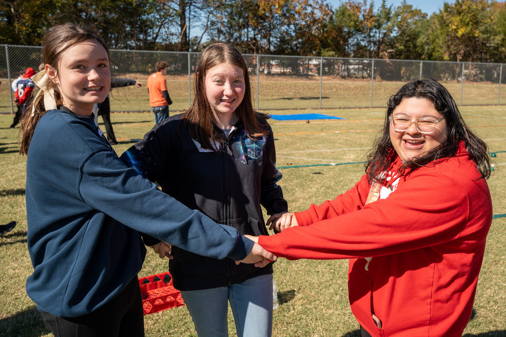 Three teenage girls stand on the grassy field holding hands in a circle. The girl on the right in a red sweatshirt is laughing joyfully.