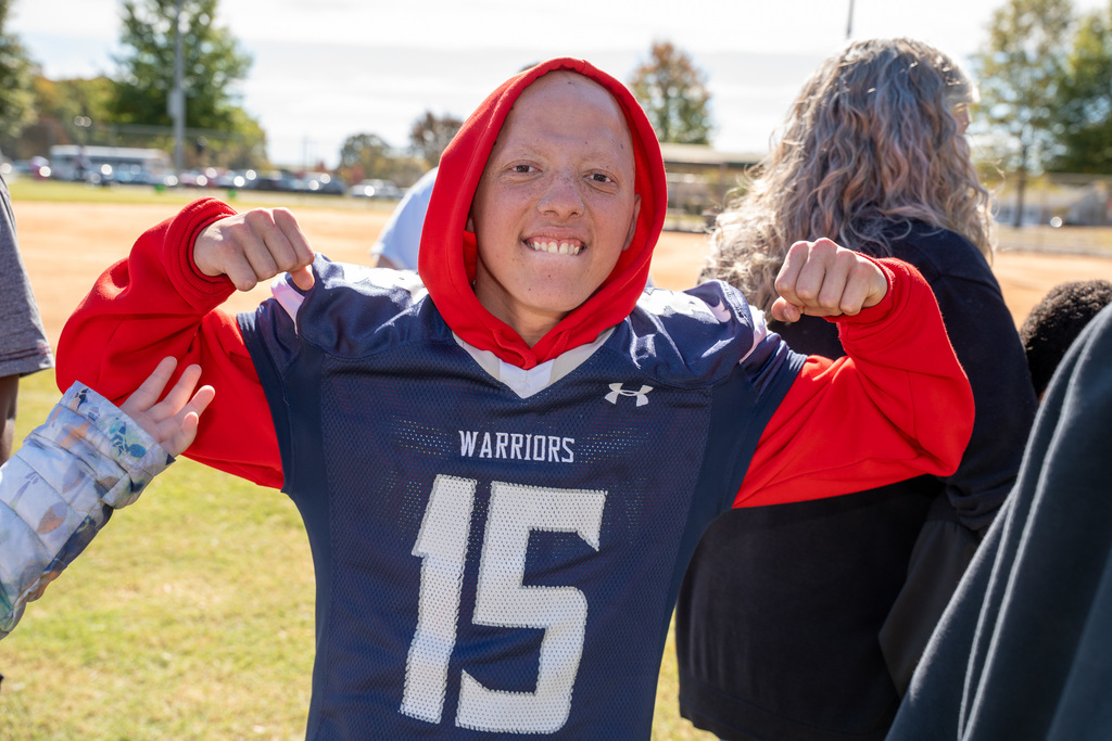 A young man in a blue and red "Warriors 15" football jersey with his red hood up smiles widely and flexes his biceps at the camera, looking strong and happy.