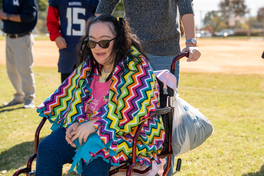 A young woman wearing sunglasses and wrapped in a vibrant, multi-colored zigzag blanket sits in a wheelchair. A person stands behind her, pushing the chair.
