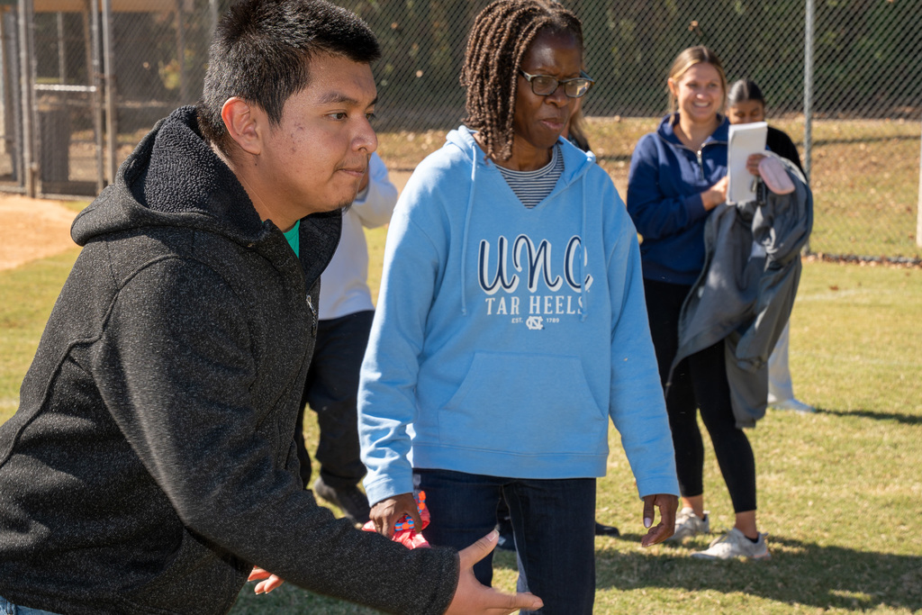 A young man in a black hooded sweatshirt leans forward, reaching out with his hands as if rolling a ball for a game. An adult woman in a light blue UNC hoodie stands behind him, watching.