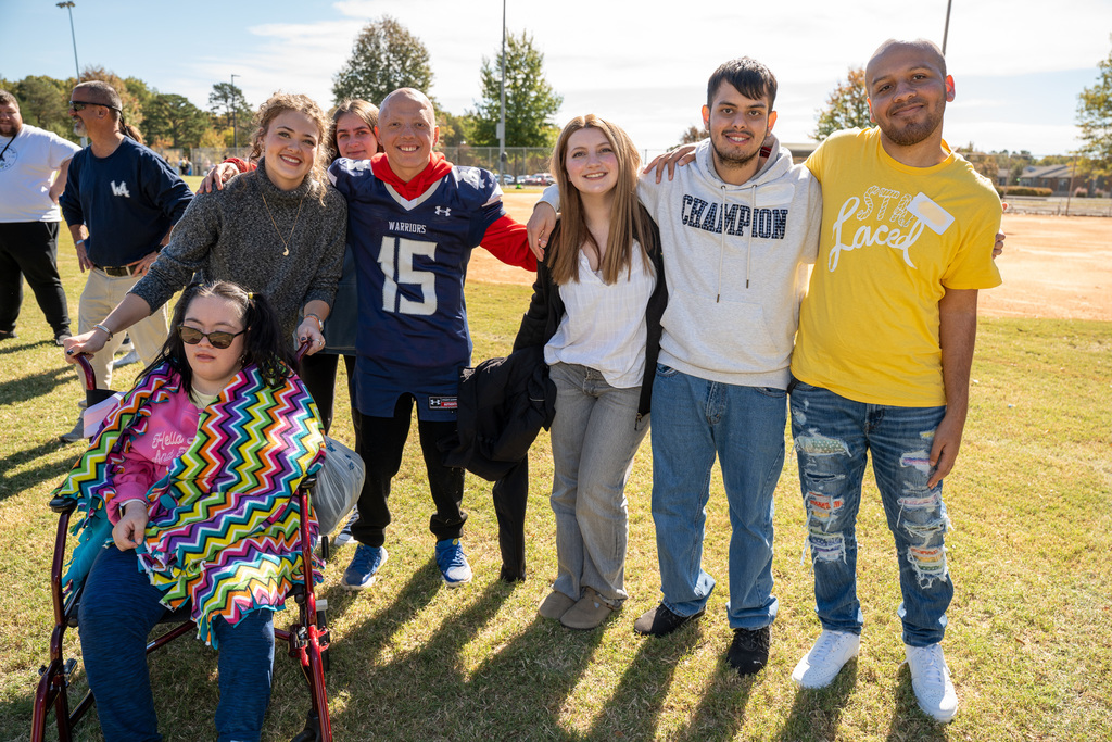 A group of six young adults and teens, including participants and volunteers, pose together for a group photo on the grassy field. One girl sits in a wheelchair in the foreground, wrapped in a colorful blanket.
