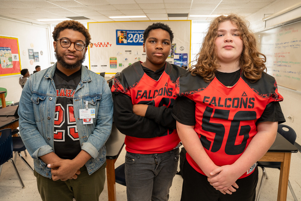 A teacher and two male students standing side-by-side in a classroom. The students are wearing red and black Graham Falcons football jerseys.