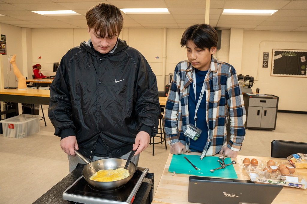 Two male students in the Career Lab cooking an omelet. One student is carefully stirring eggs in a small skillet on a portable burner, while the other watches with eggs and cooking tools on the counter.
