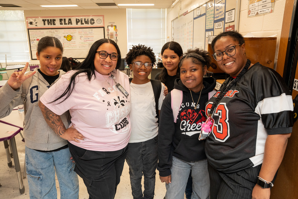 A group photo of four students and two teachers in a brightly lit classroom. Two students are flashing peace signs, and the adults are smiling warmly.