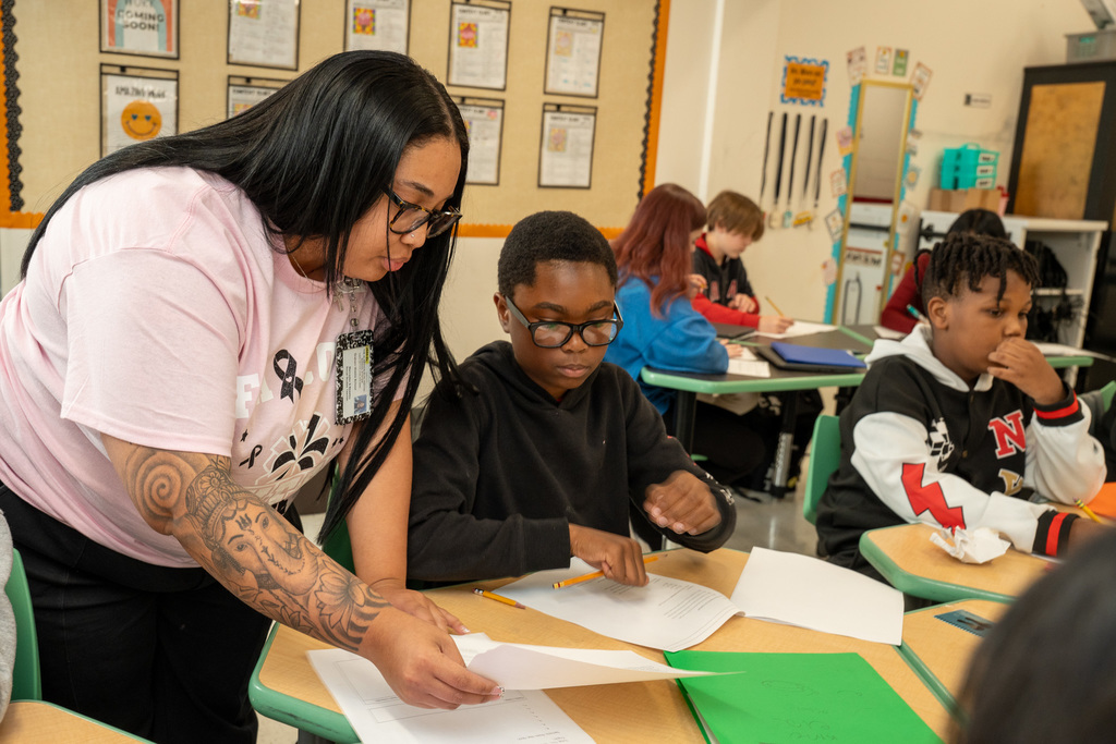 A teacher with long dark hair and glasses leaning down to help a male student at his desk, pointing to a paper while he holds a pencil. Other students are working in the background.