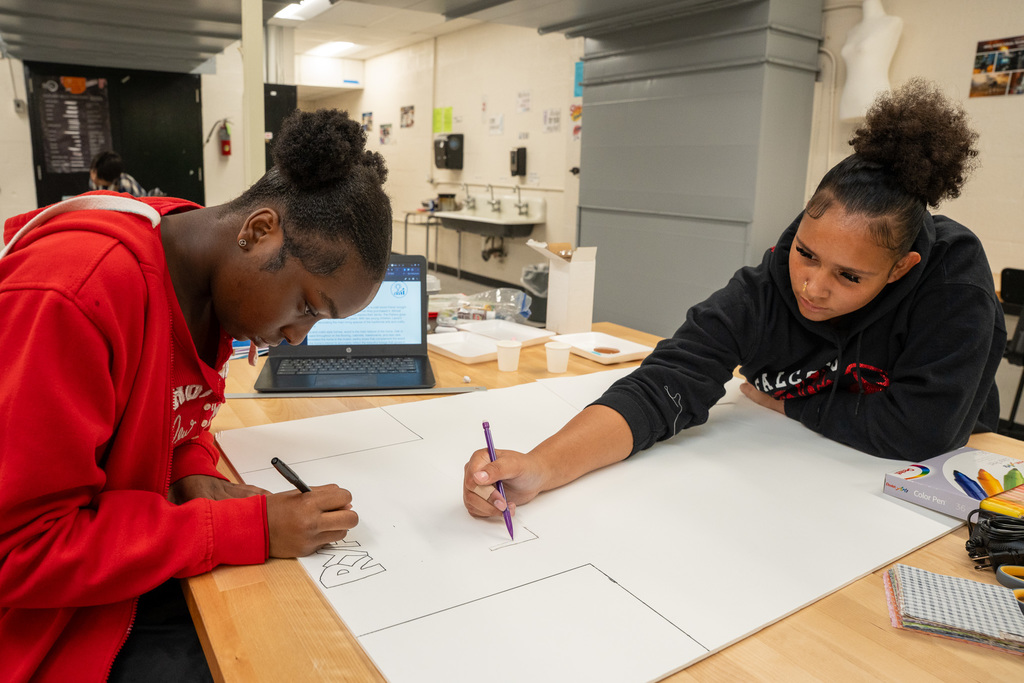 Two female students intently working on a large piece of poster board in the Career Lab, using markers and pencils to draw a design. A laptop is open in the background.