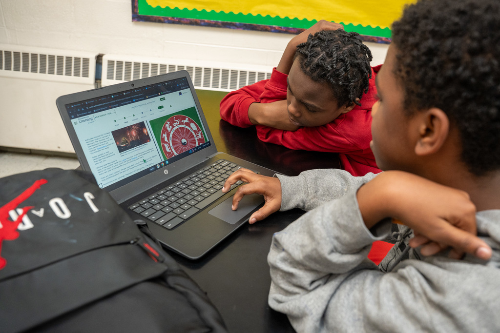 Two male students in a classroom looking at a laptop screen. One student is resting his head on his arms, and the other is navigating a website with the touchpad.