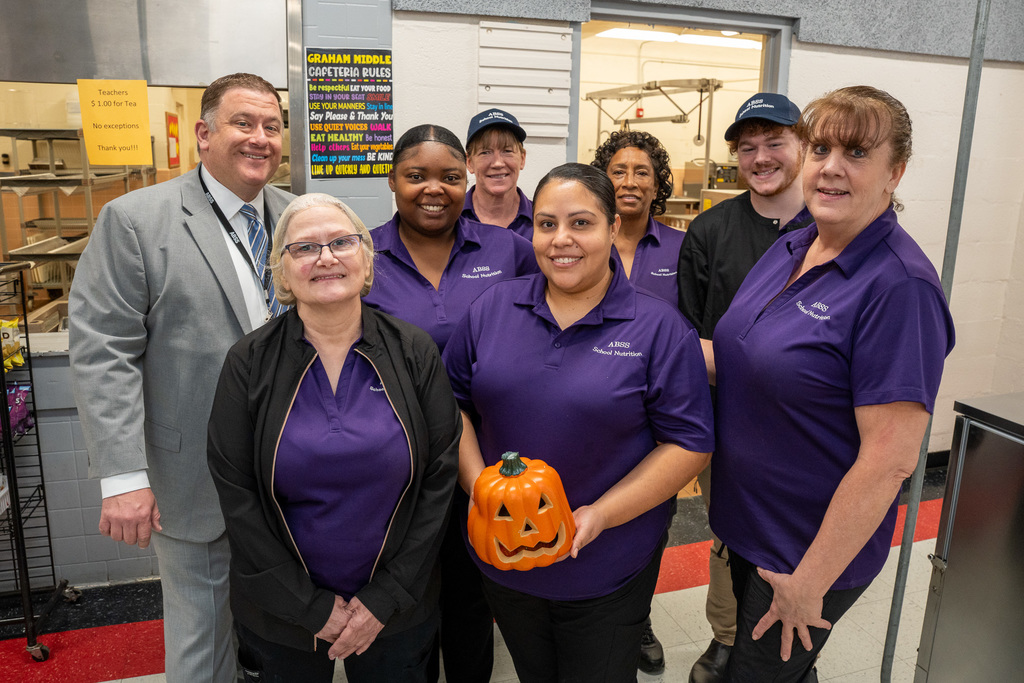 A group of seven friendly Graham Middle School cafeteria staff in purple shirts, along with a man in a gray suit (Dr. Fleming), posing for a photo in the kitchen area. One staff member in the center is holding a carved orange jack-o'-lantern.