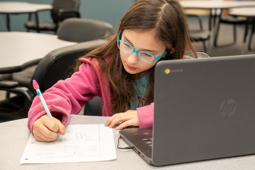 A female student with long hair and bright blue glasses focused on writing notes on a sheet of paper next to her open laptop.
