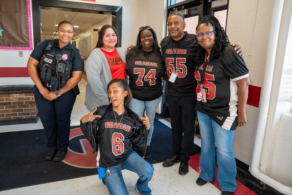A group of six smiling adults, including a School Resource Officer (SRO) and several teachers wearing red and black Graham Falcons football jerseys, posing together in a hallway.