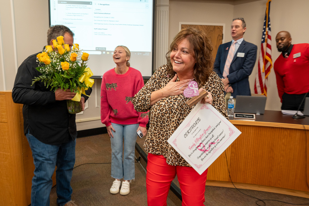 Board Chair Sandy Ellington-Graves stands surprised holding a certificate recognizing her as a breast cancer survivor.  Her daughter and husband stand behind her with exuberant expressions.
