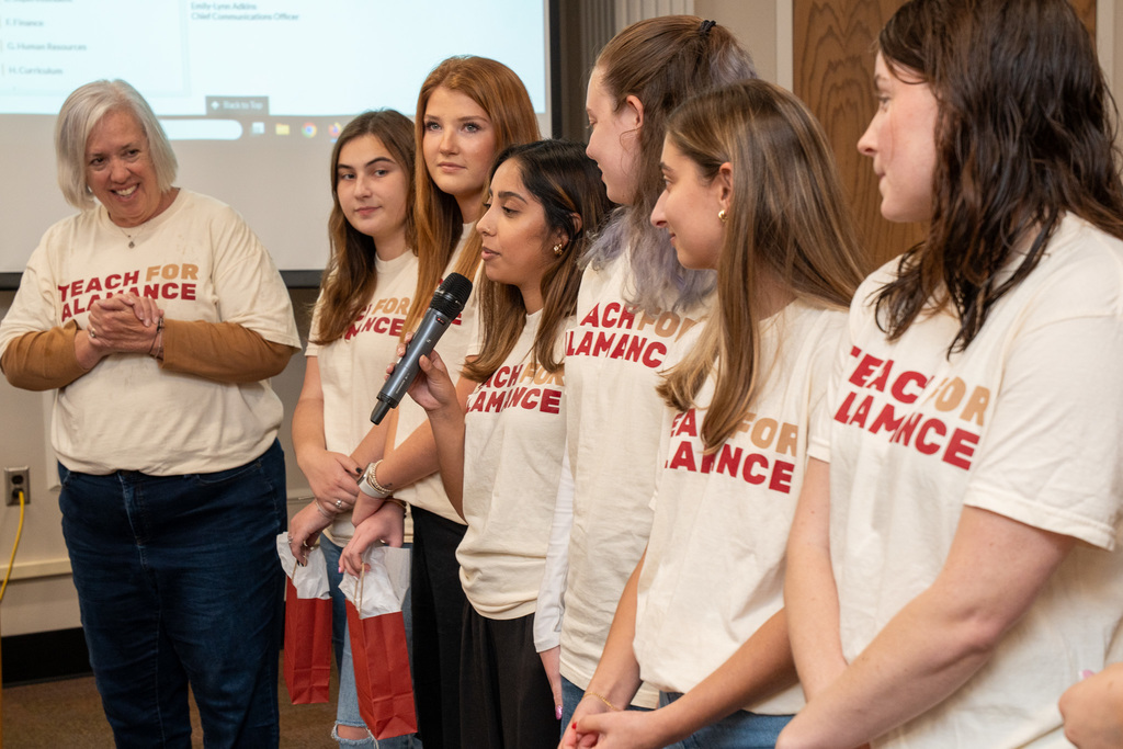 A close-up shot of five women in the Teach for Alamance cohort, with one woman speaking into a microphone. The woman with short grey hair stands to the far left, clapping.