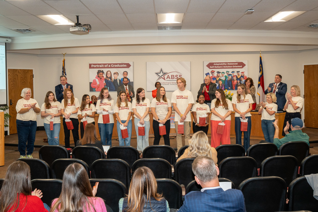 A wide shot of the Teach for Alamance cohort standing and being applauded by several adults on either side and the audience in the foreground. They are standing in front of large ABSS posters.