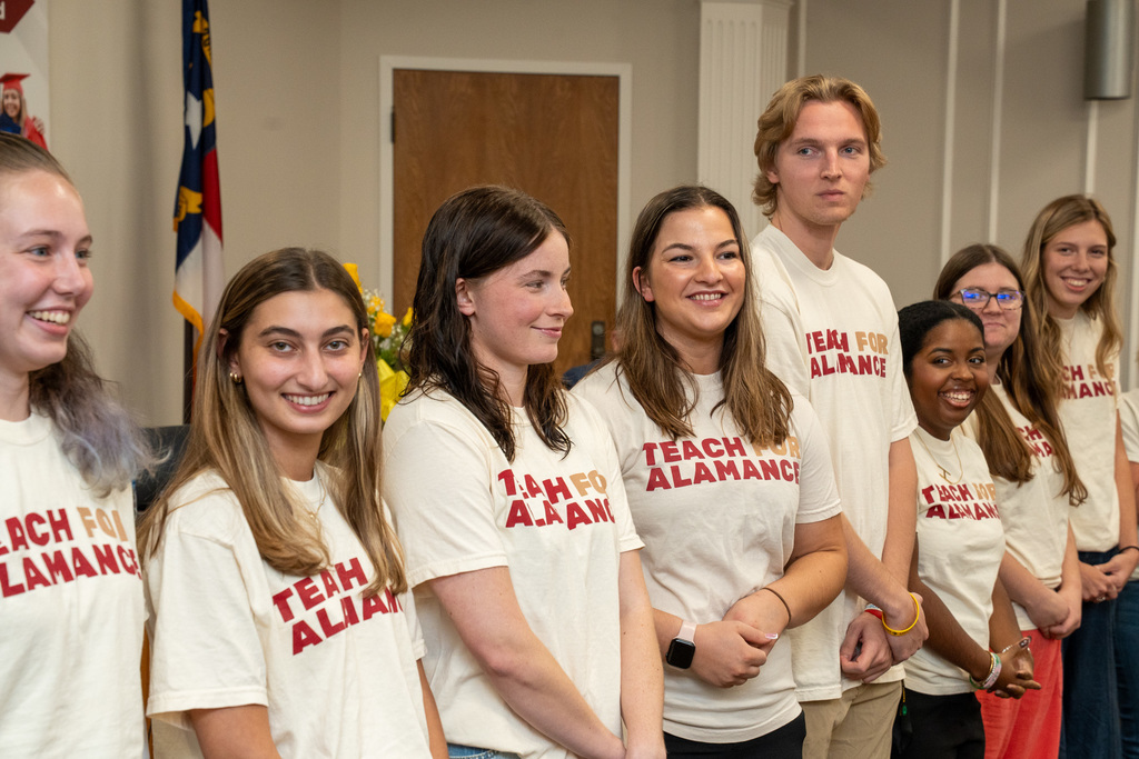 A close-up side view of six members of the Teach for Alamance cohort, smiling and standing in a row in their matching t-shirts.