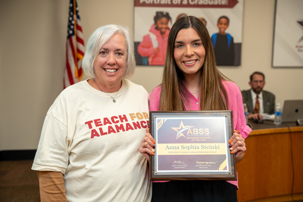 A woman with short grey hair (Dean Ann Bullock) wearing a "TEACH FOR ALAMANCE" shirt stands next to a young woman (Anna Steinki) wearing a pink shirt. The young woman holds a framed certificate that reads "ABSS BEGINNING TEACHER OF THE YEAR 2024-2025: Anna Sophia Steinki." Both are smiling for the camera.