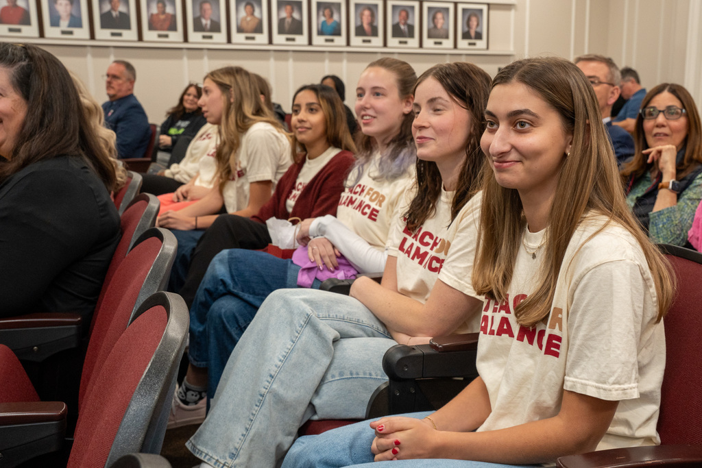 A side view of four young women from the Teach for Alamance cohort sitting and listening in the audience of the meeting room. They are wearing their matching t-shirts.
