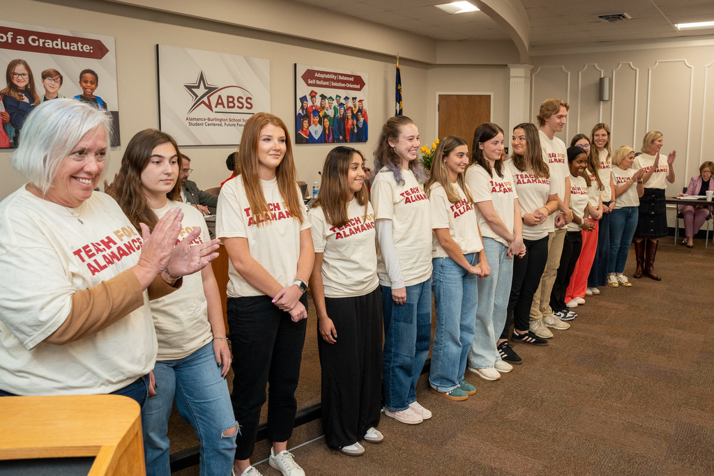 A group of young adults, mostly women, wearing matching light-colored t-shirts that say "TEACH FOR ALAMANCE" in red lettering, are standing in a line in a meeting room. A woman with short grey hair is clapping enthusiastically near the foreground on the left.