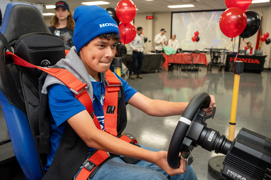 A male student wearing a blue beanie and safety harness intensely operates a realistic ATV driving simulator (MOZA Racing wheel), providing a fun, hands-on experience of vehicle operation.