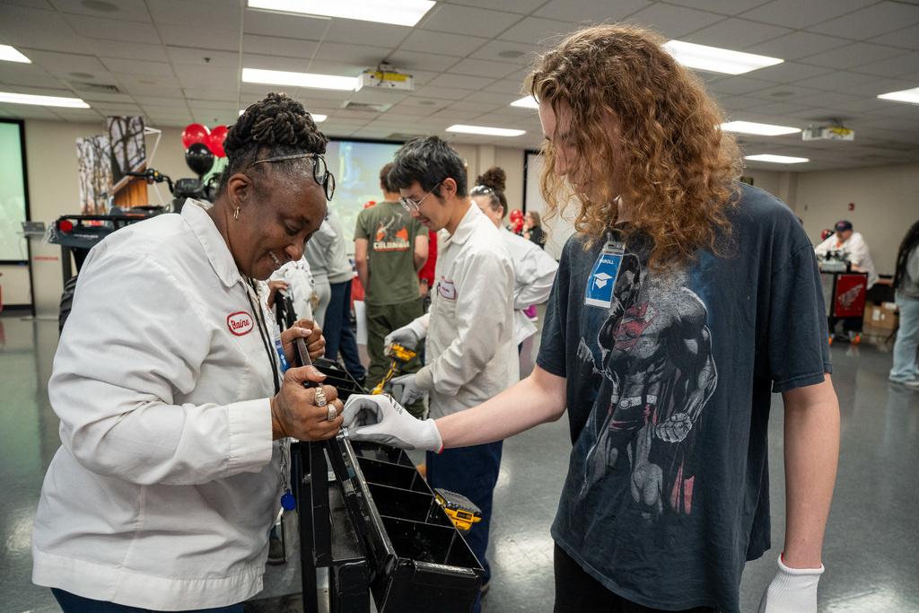 A Honda team member in a white uniform assists a male high school student with long curly hair in assembling a part on a black ATV component, demonstrating a hands-on manufacturing skill.