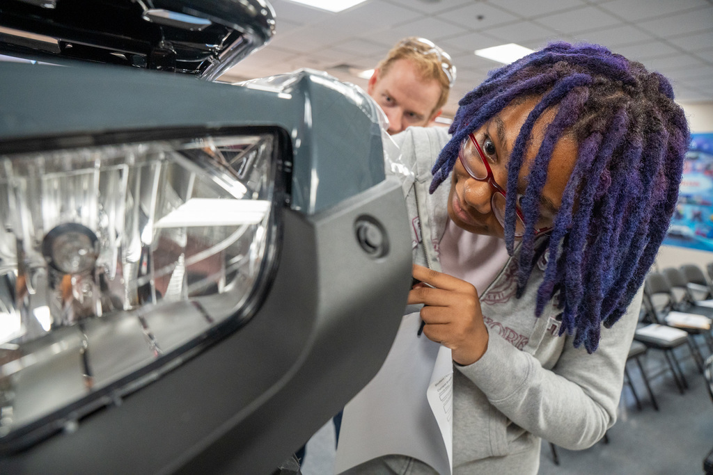 Close-up of a female student with purple dreadlocks carefully inspecting the fit and finish of a front ATV plastic fascia and headlight assembly. A Honda team member is visible behind her.