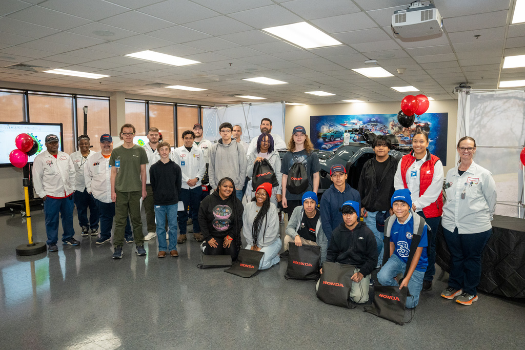 A group photo of the high school CTE students and Honda team members inside a training room. Students are kneeling in front with black Honda bags, celebrating the completion of their manufacturing day tour.