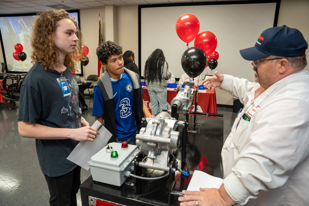 A Honda team member (wearing a hat) points to and explains the workings of a mechanical engine component display to two male students, demonstrating the technical details of the ATV powertrain.