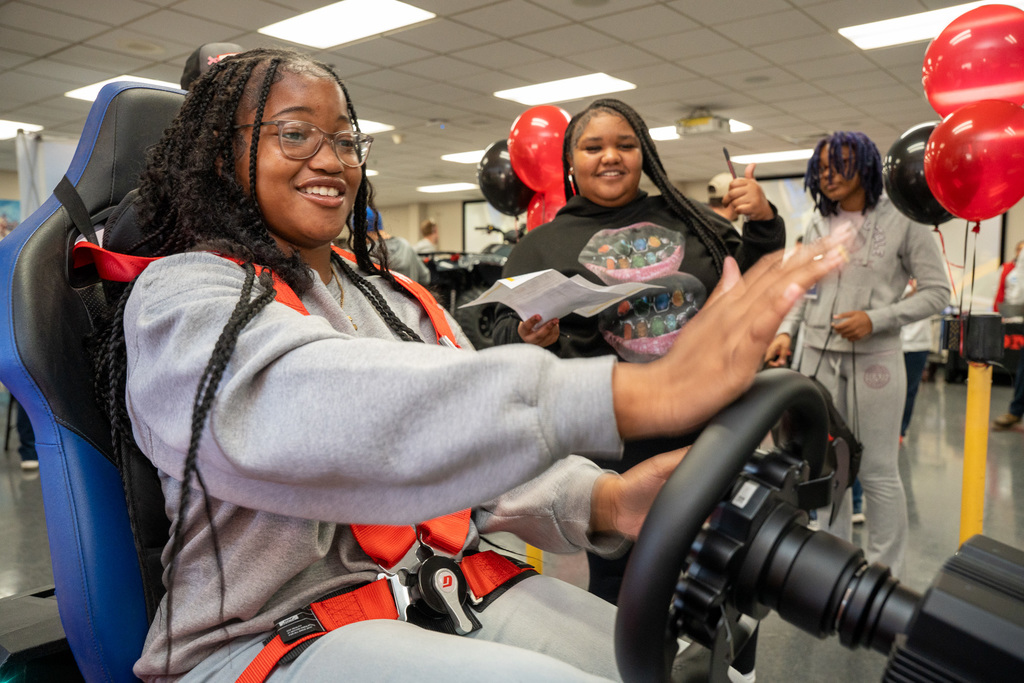 A female student in a gray sweatshirt smiles enthusiastically while operating the ATV driving simulator. Another female student watches and gives a thumbs-up in the background.