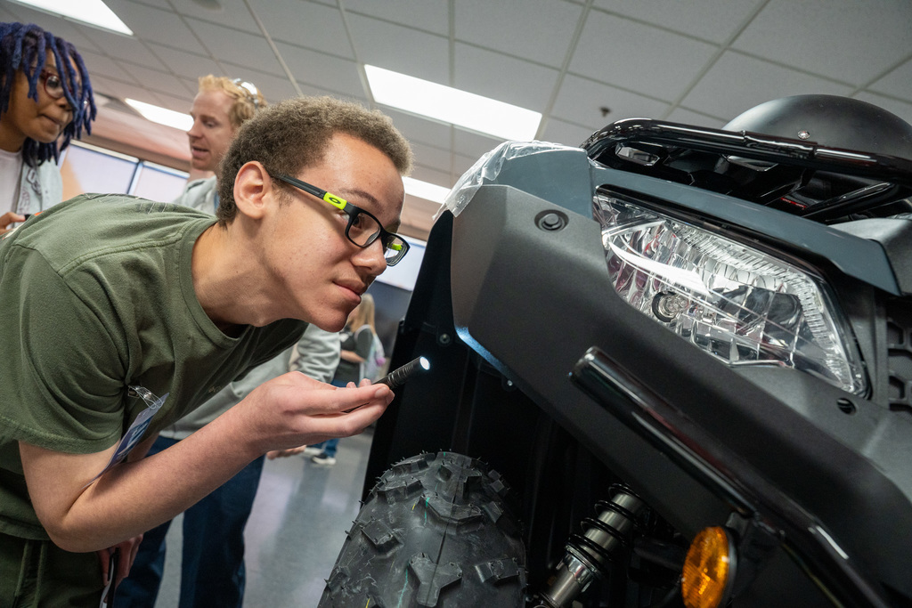 A male student wearing glasses and a green shirt leans in close, inspecting the front headlight and tire of an ATV model with a small penlight, focusing on quality and assembly detail.